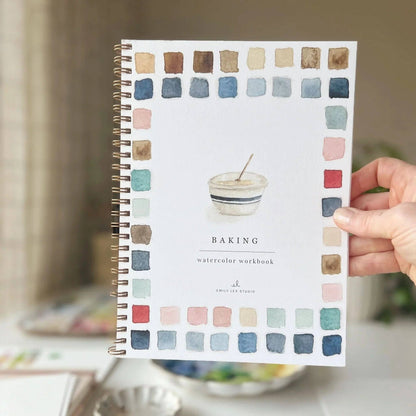 Hand holding a baking watercolor workbook with color swatches on the cover.