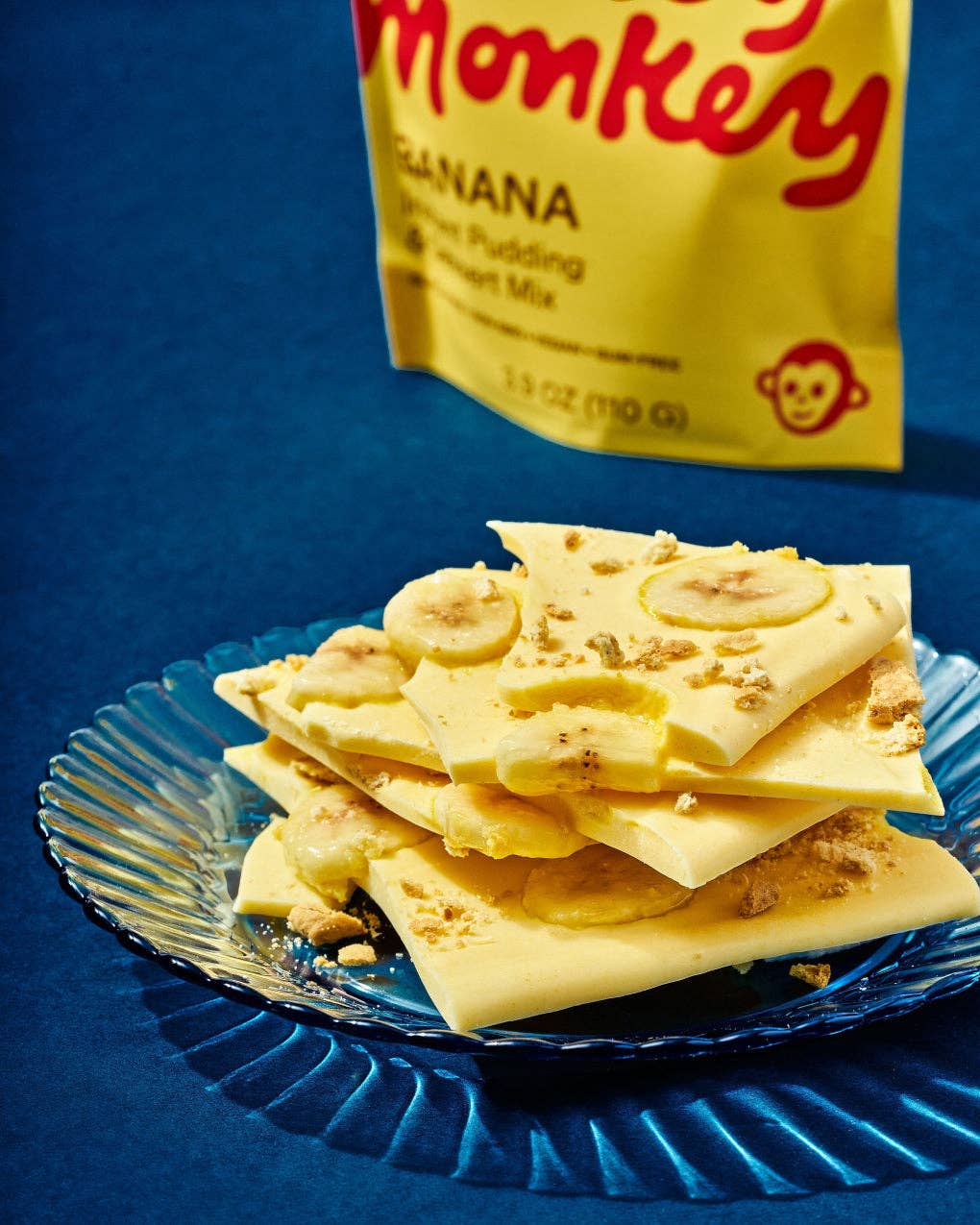 Banana pudding bark topped with banana slices and crumbled peanuts, served on a blue scalloped plate with a yellow bag of Sweet Monkey Banana Pudding in the background.