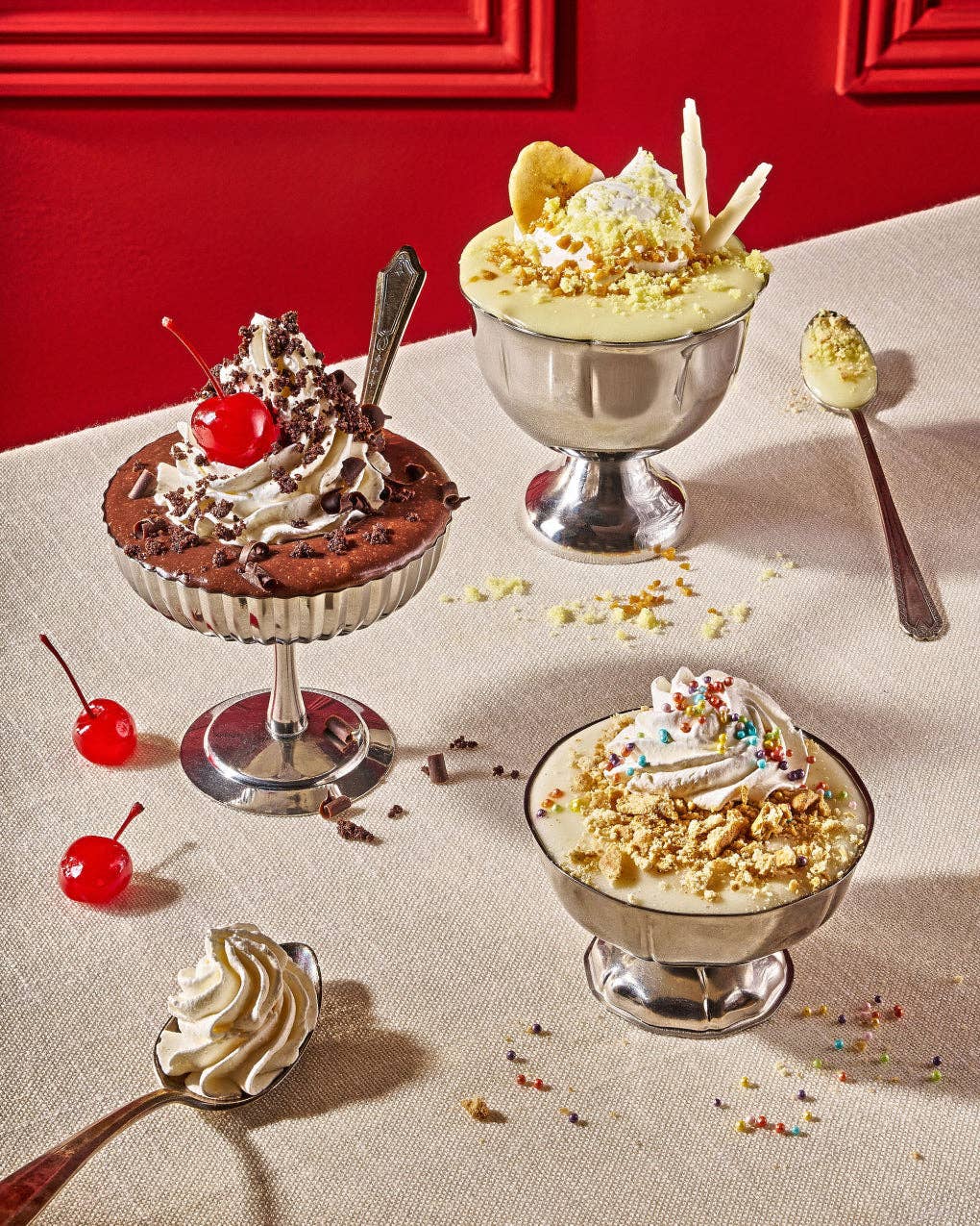 Three dessert parfaits in silver pedestal bowls arranged on a beige tablecloth with a red wall background. The desserts include whipped cream, crushed nuts, sprinkles, chocolate pieces, and cherries, with additional whipped cream and nuts placed on spoons nearby.