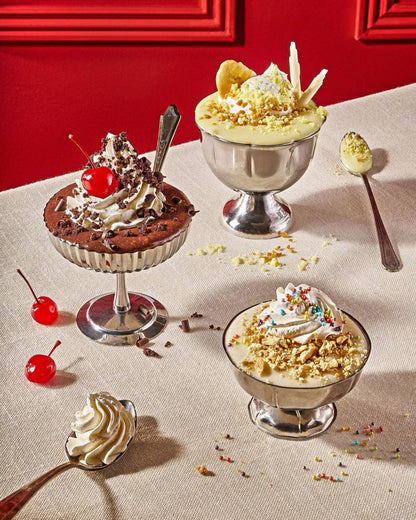Three dessert parfaits in silver pedestal bowls arranged on a beige tablecloth with a red wall background. The desserts include whipped cream, crushed nuts, sprinkles, chocolate pieces, and cherries, with additional whipped cream and nuts placed on spoons nearby.