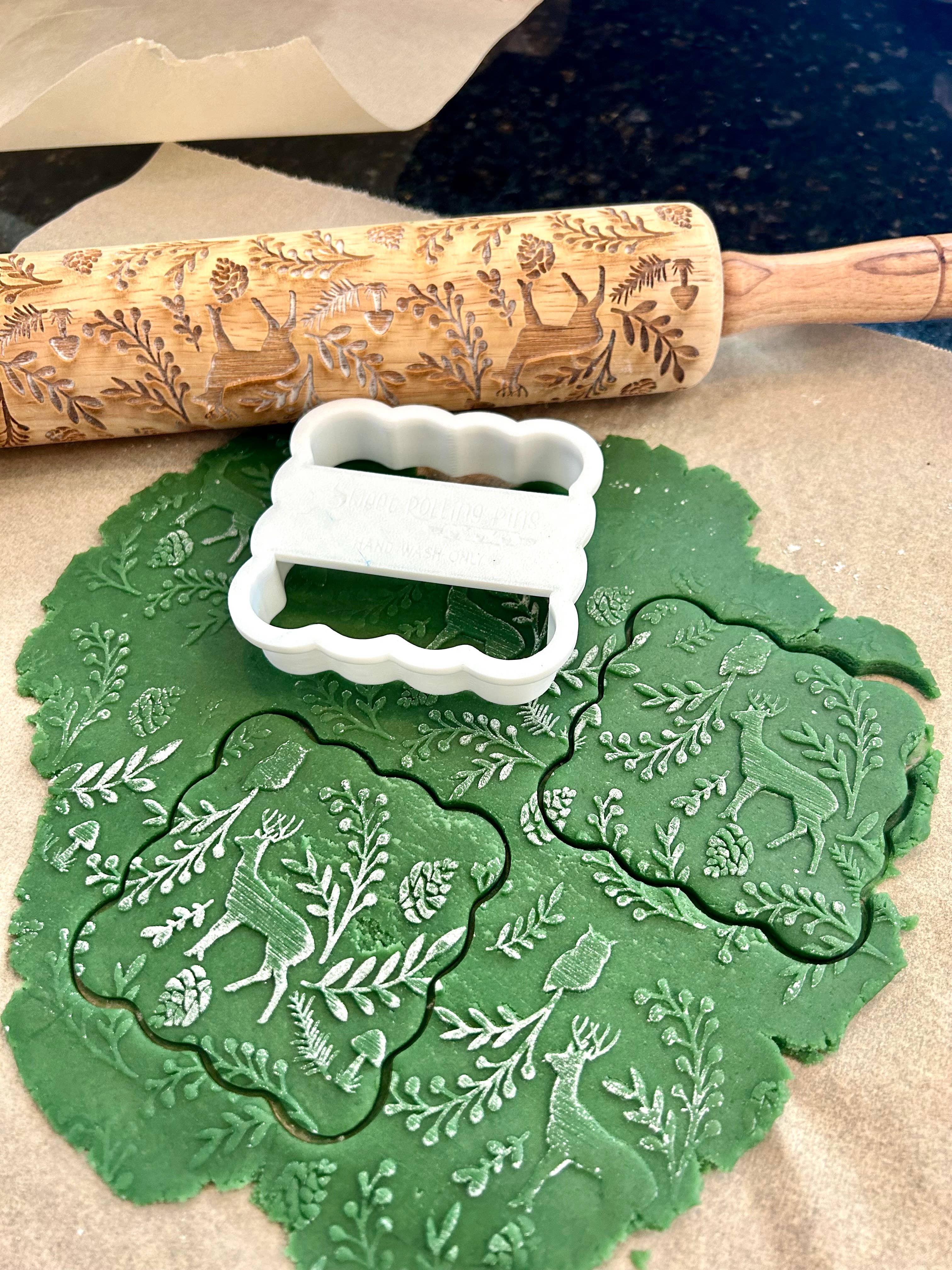 A white scalloped cookie cutter with a festive woodland design sits on top of green rolled-out dough on parchment paper, with a wooden rolling pin engraved with similar motifs in the background.