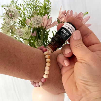 Hand holding a bottle of essential oil with a beaded bracelet and flowers in the background