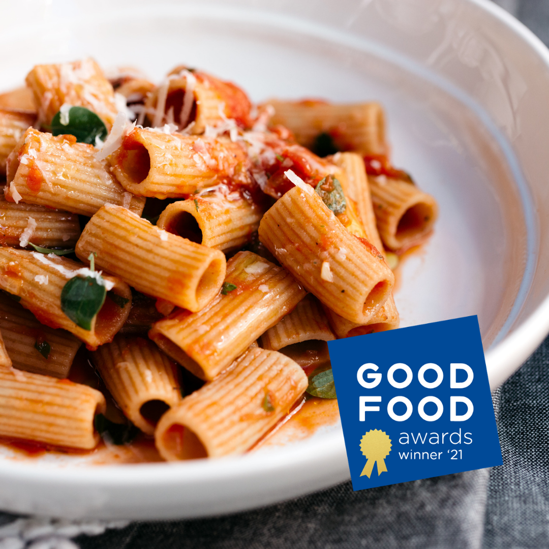 A close-up of a white bowl filled with rigatoni pasta in a red tomato sauce, topped with grated cheese and basil leaves. A blue &