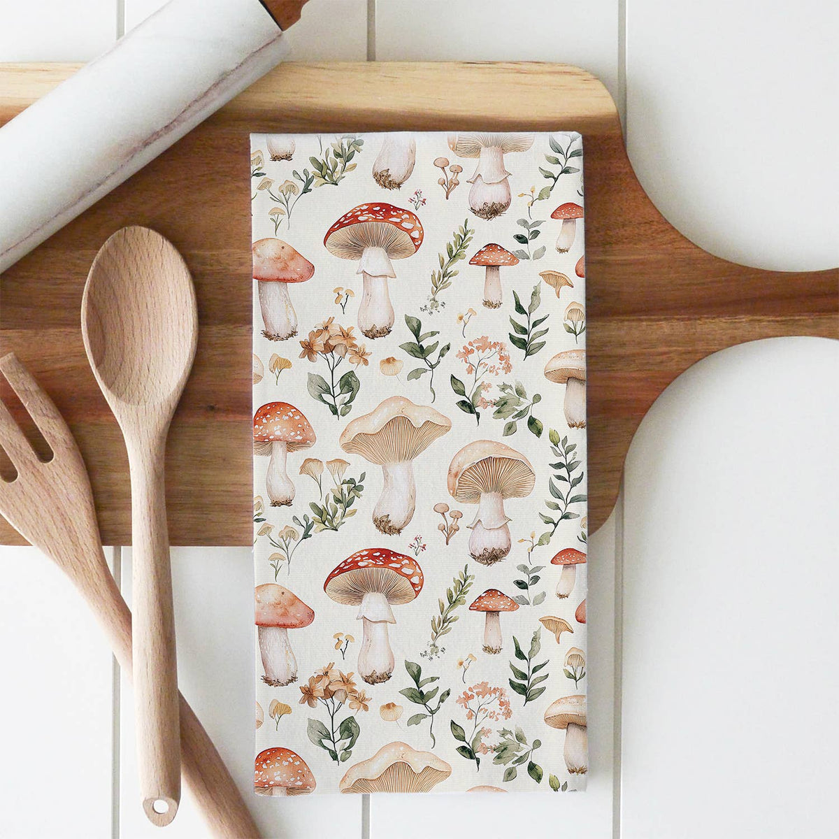 Kitchen towel with mushroom and floral pattern on a wooden cutting board with utensils.