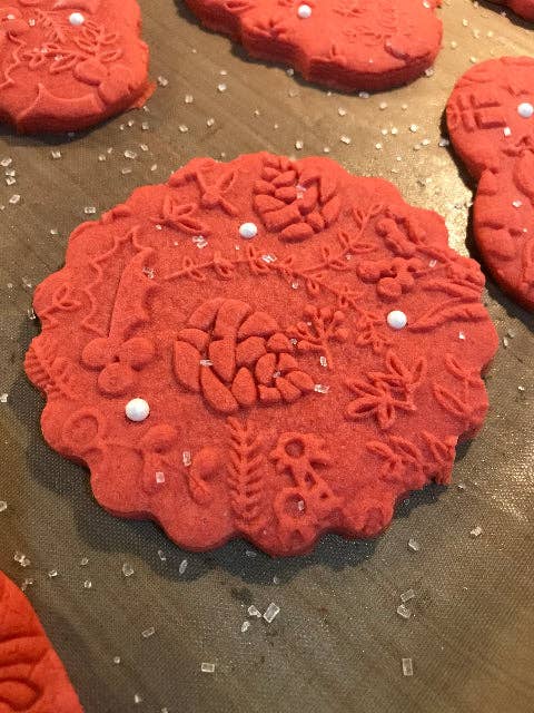A close-up of a round red decorated cookie with embossed floral and geometric patterns, topped with small white pearl sprinkles, on a dark textured surface.