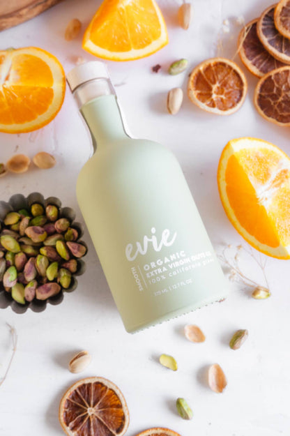 Bottle of Evie organic extra virgin olive oil with sliced oranges and pistachios on a white surface