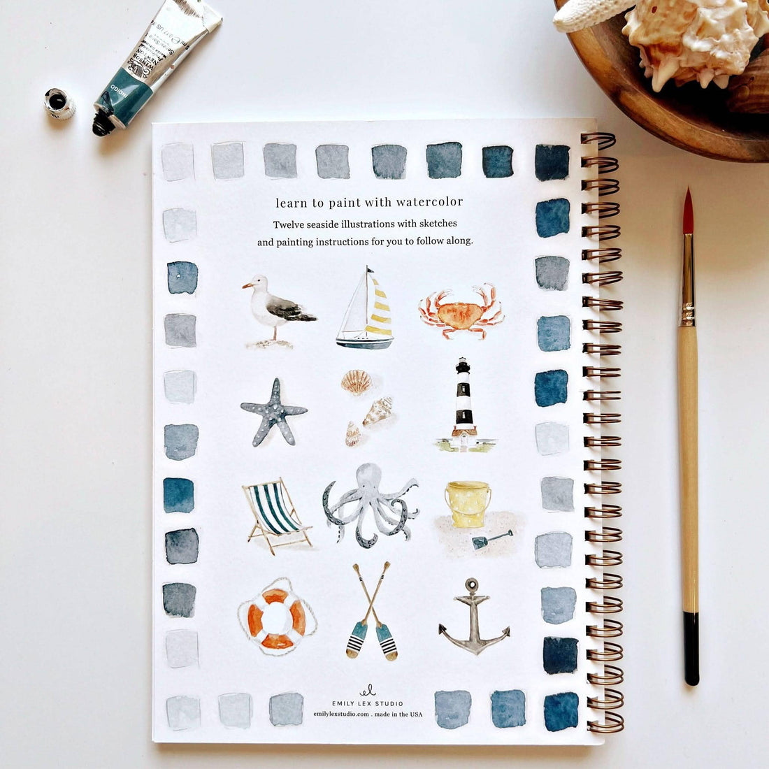 Notebook with watercolor illustrations of sea-themed objects on a white surface with a paintbrush and seashells.