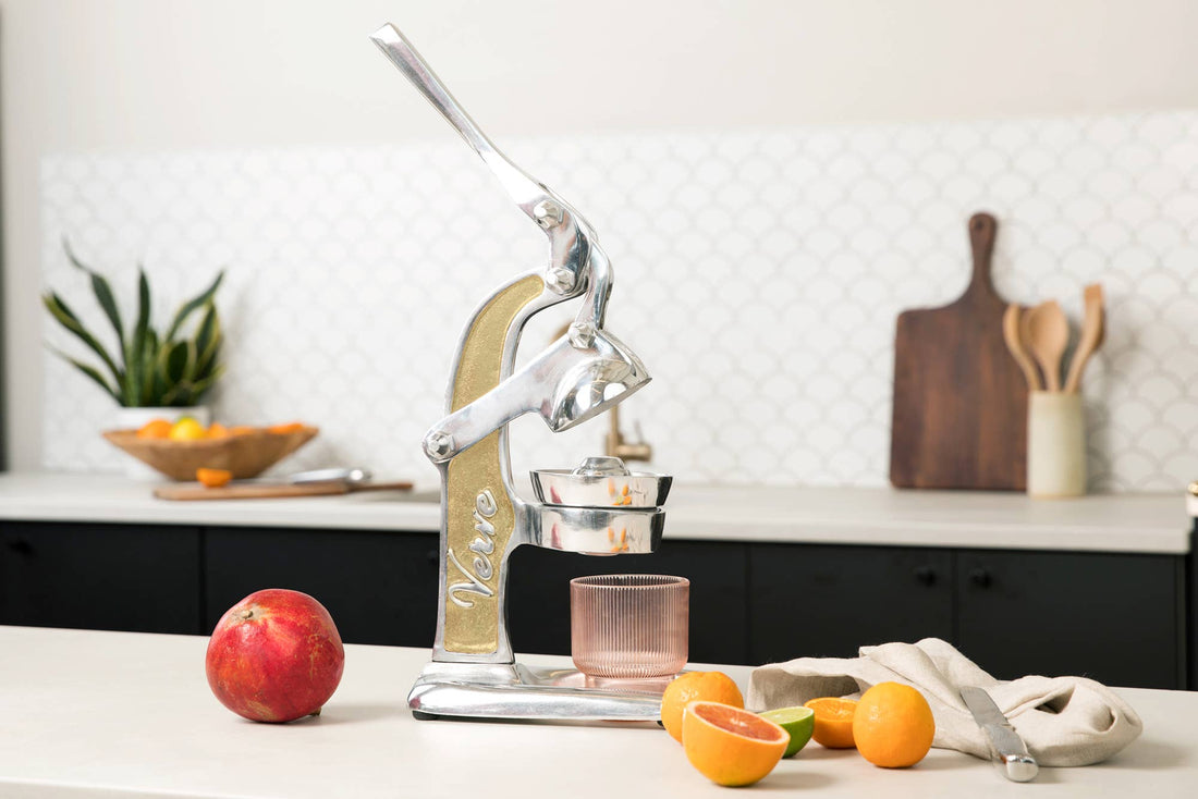Manual juicer on a kitchen counter with fruits and a glass.