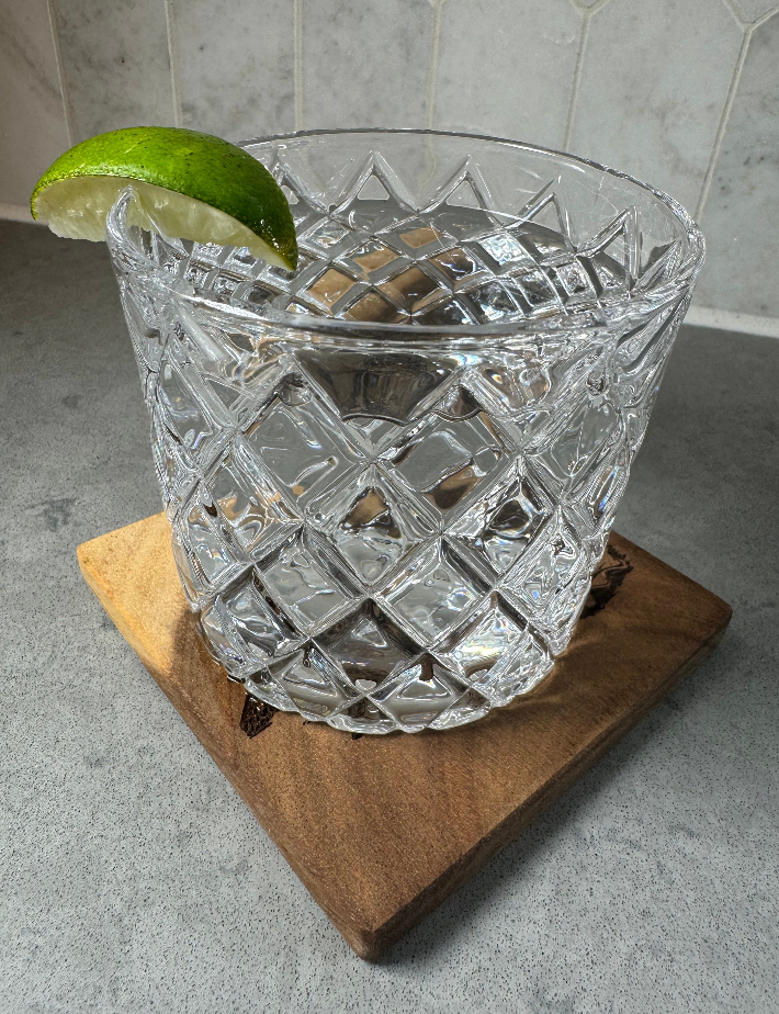 A clear cut-crystal tumbler with a diamond pattern, filled with ice and garnished with a lime wedge, resting on a wooden board against a gray tiled background.