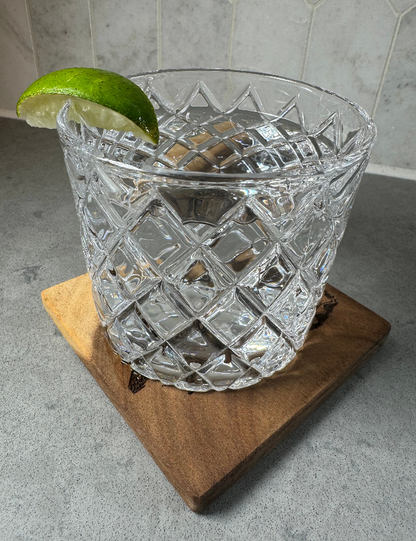 A clear cut-crystal tumbler with a diamond pattern, filled with ice and garnished with a lime wedge, resting on a wooden board against a gray tiled background.