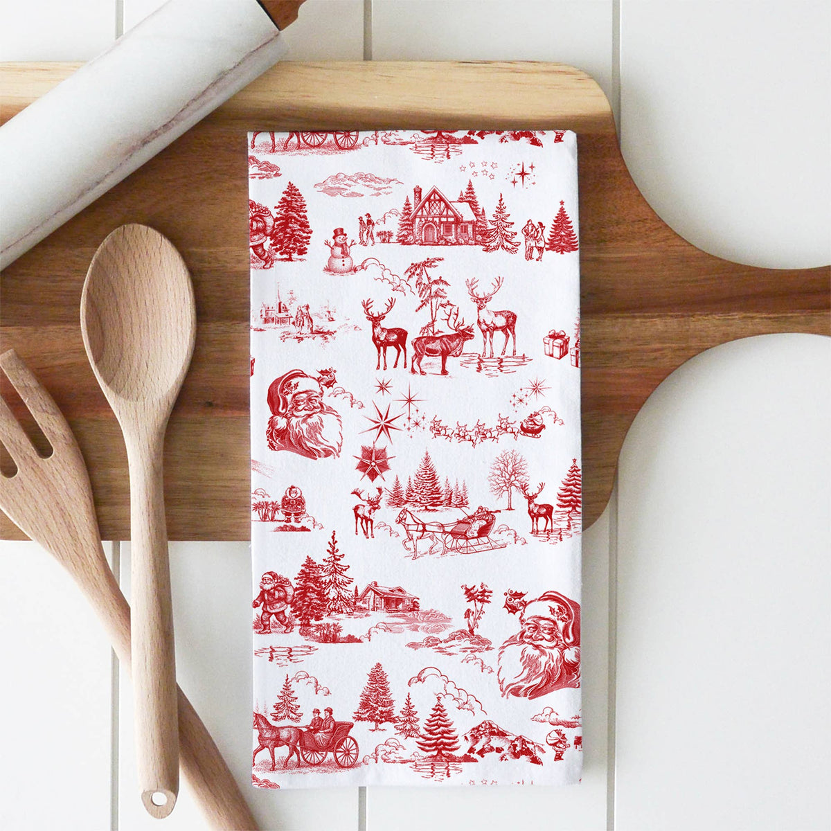 A white kitchen tea towel with a red Christmas toile pattern, laid flat on a wooden cutting board with wooden spoons and a rolling pin as props.