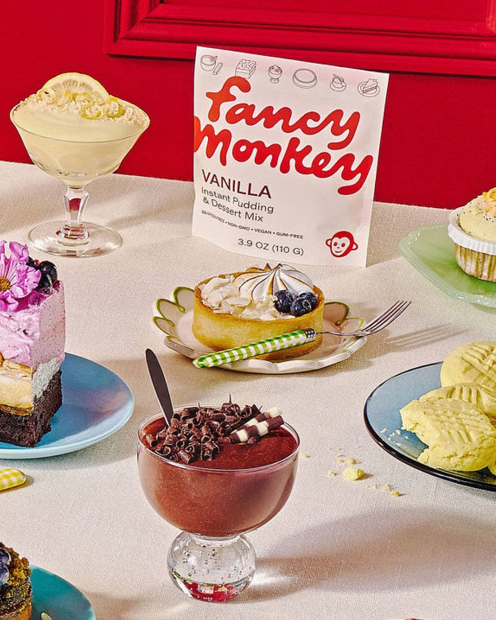 A package of Fancy Monkey Vanilla Ice Cream Pudding &amp; Dessert Mix stands on a table surrounded by styled desserts, including a vanilla pudding cupcake, chocolate pudding with whipped cream and chocolate shavings, a pink layered cake, and yellow cookies, against a red wall background.