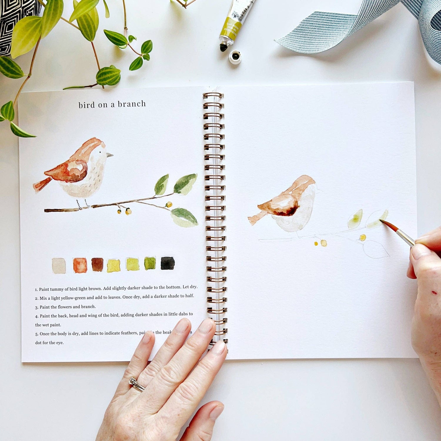 Artistic depiction of a bird on a branch with watercolor painting supplies and a color swatch.