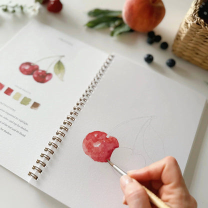 Person painting a cherry on a sketchbook with fruits and a basket in the background