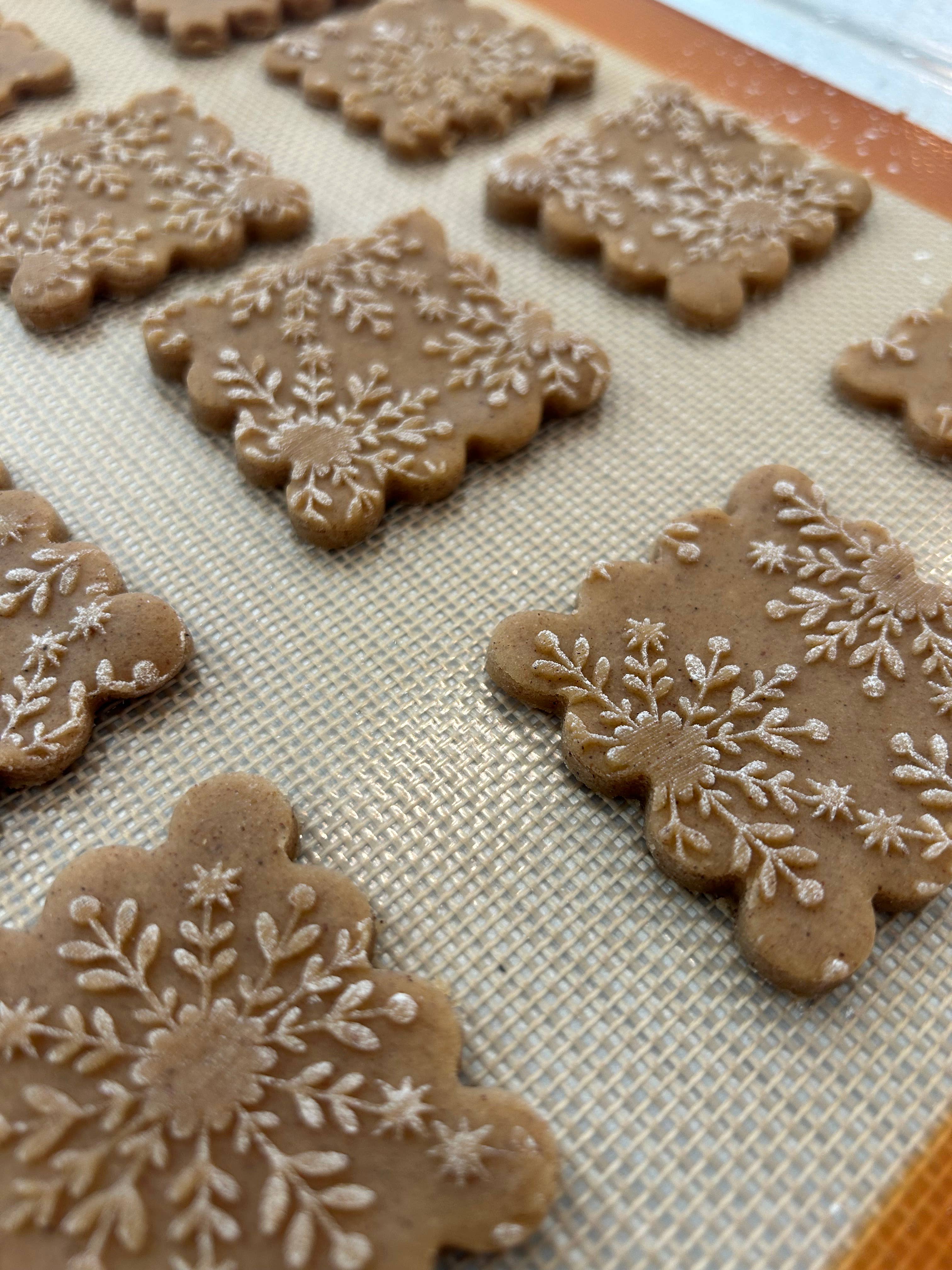 Decorated gingerbread cookies with intricate white icing snowflake patterns, arranged on a beige textured silicone baking mat.