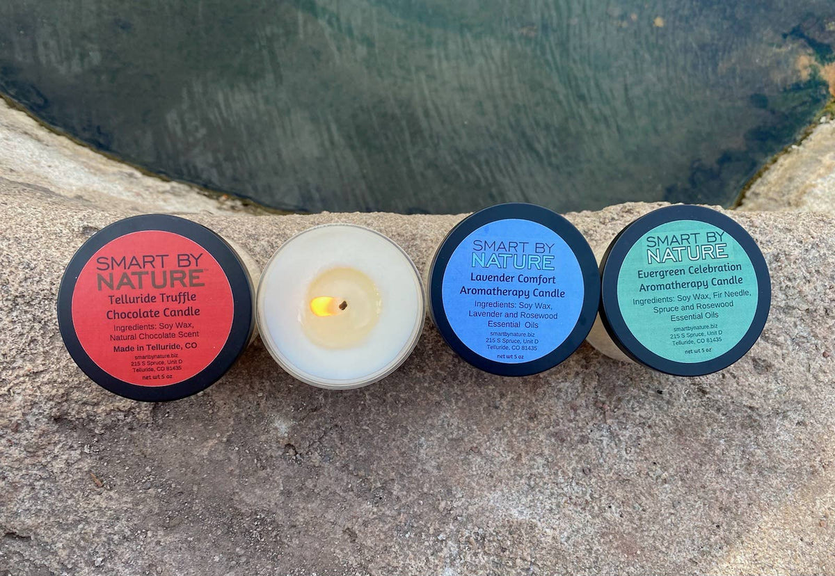 Four Smart By Nature candles are lined up on a stone surface with a dark, rocky background. From left to right: a Telluride Truffle Chocolate Candle with a red label, a white wax candle with a lit flame, a Lavender Comfort Aromatherapy Candle with a blue label, and a Green Celebration Aromatherapy Candle with a green label.
