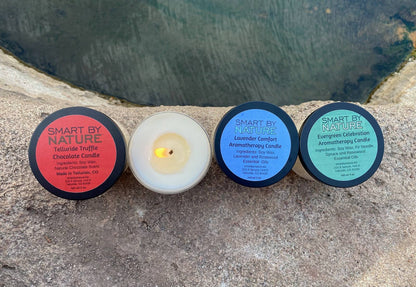 Four Smart By Nature candles are lined up on a stone surface with a dark, rocky background. From left to right: a Telluride Truffle Chocolate Candle with a red label, a white wax candle with a lit flame, a Lavender Comfort Aromatherapy Candle with a blue label, and a Green Celebration Aromatherapy Candle with a green label.