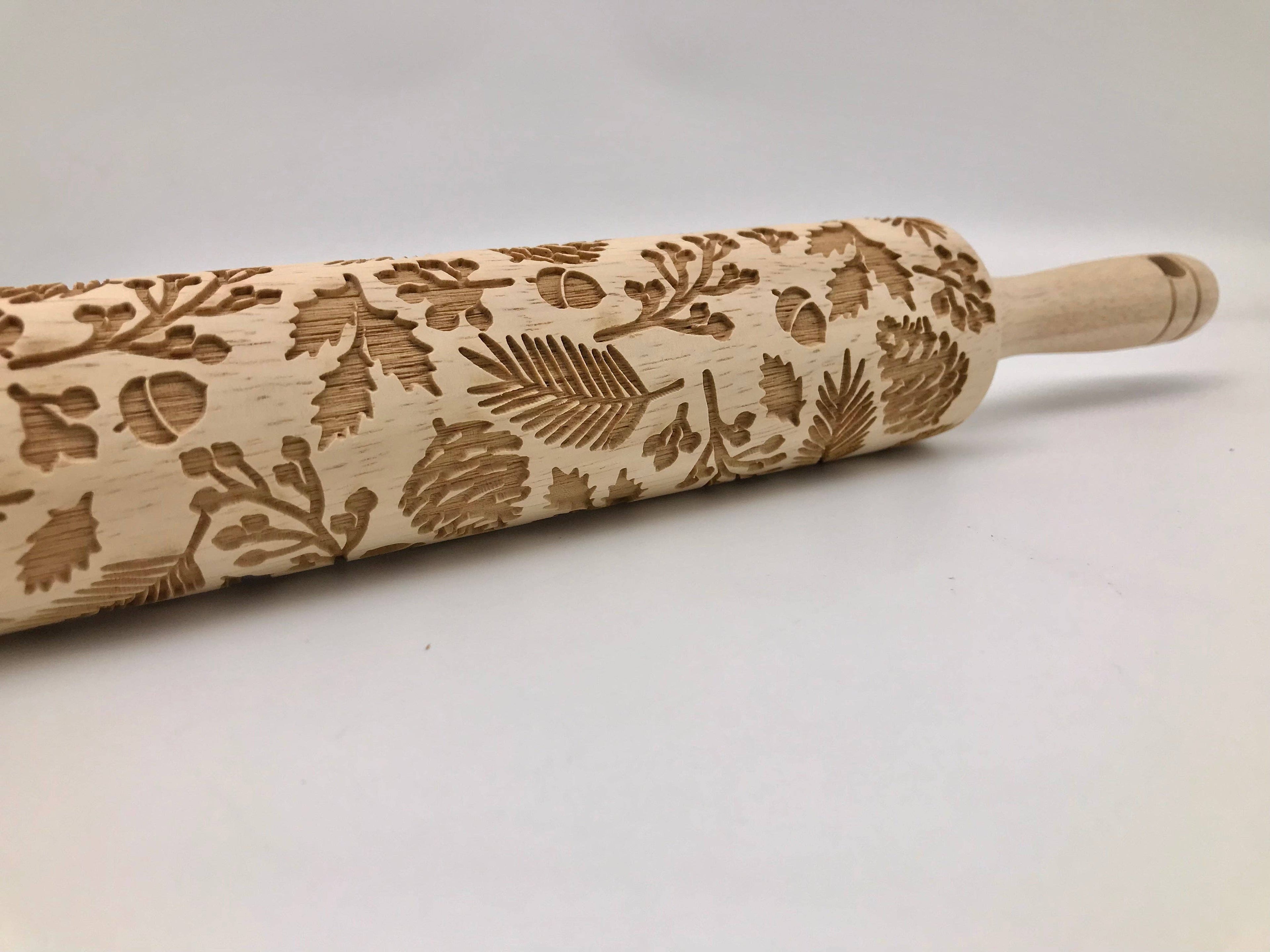 A wooden rolling pin with an engraved pattern of pine needles, pinecones, and leaves, shown on a plain light gray background.