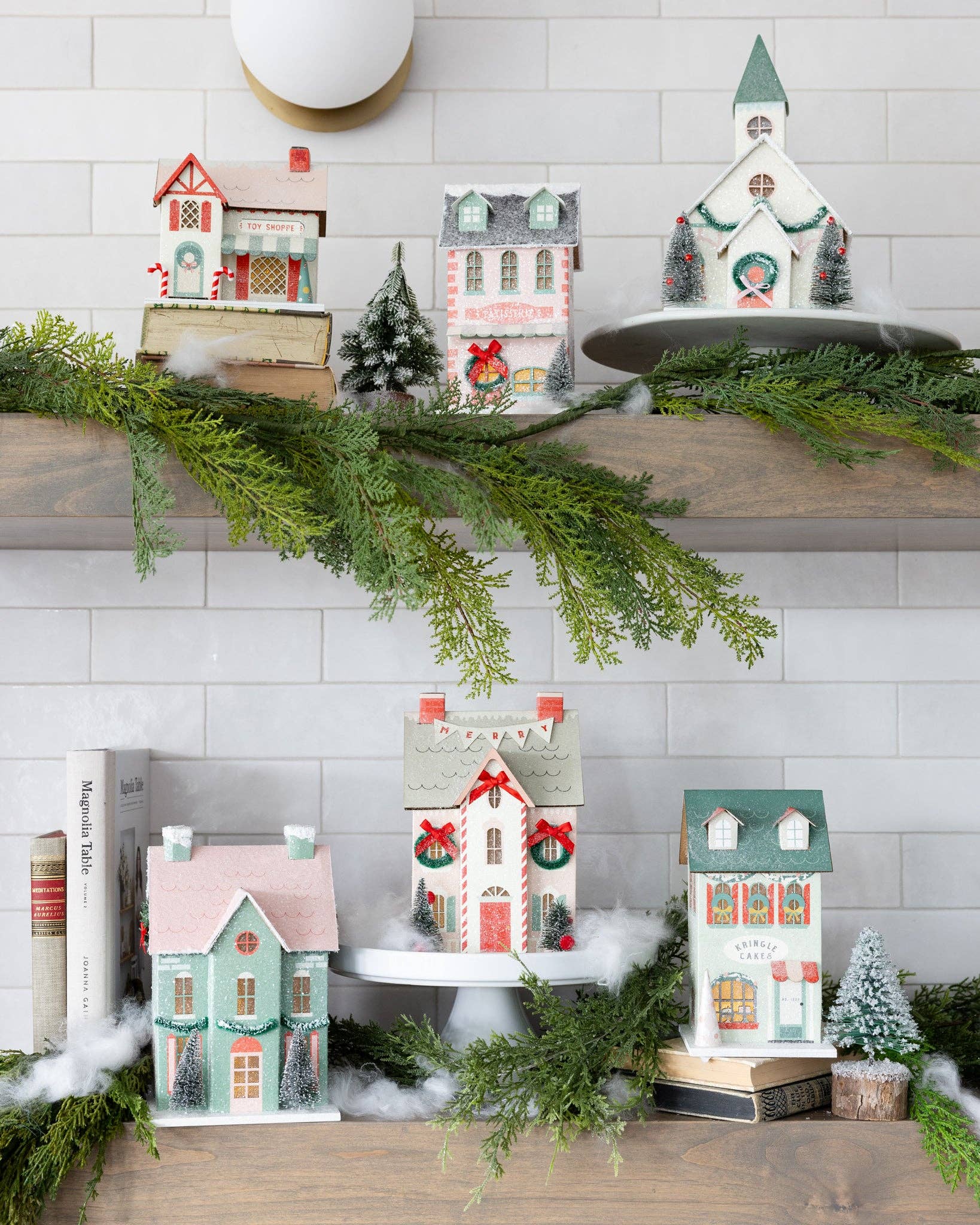 Decorative Christmas village houses on a mantel with greenery and a white wall background.