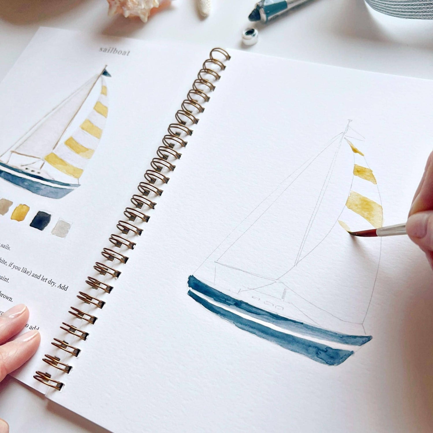 Person painting a sailboat illustration on a white surface with a paintbrush.