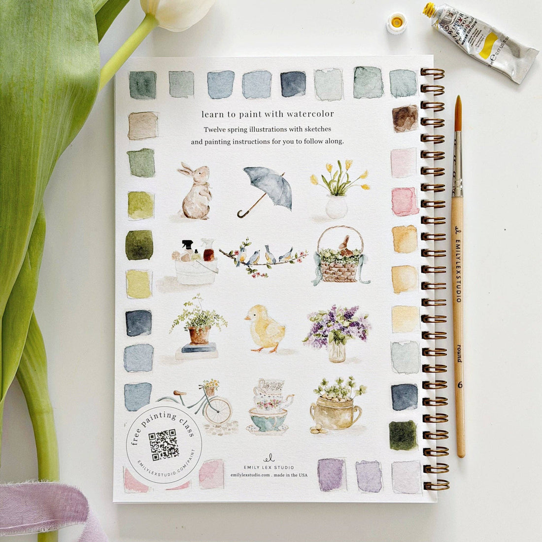 Art book with watercolor illustrations on a white surface with a plant and paint bottle.