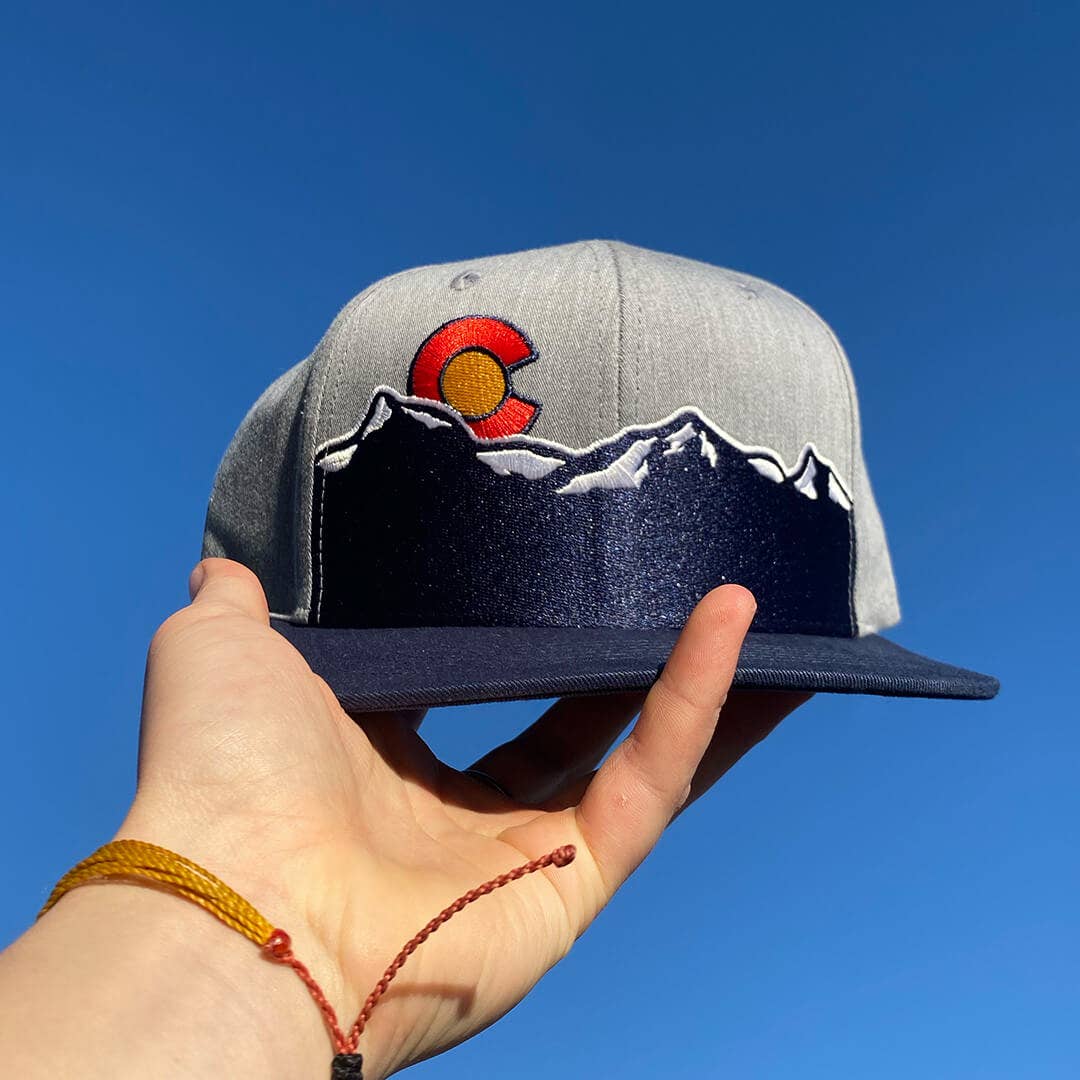 Gray cap with mountain design and red circle logo held against a blue sky