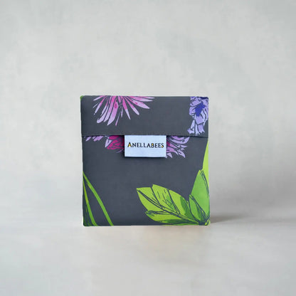 A square fabric pouch with a dark gray background and a tropical floral print in purple, pink, and green, featuring a white label with the brand name &