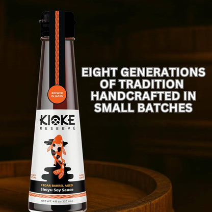 A bottle of Kioke Reserve Cedar Barrel Aged Shoyu Soy Sauce stands on a wooden barrel surface against a dark wood background. The bottle has a black cap, a white label with a koi fish illustration, and orange accents.