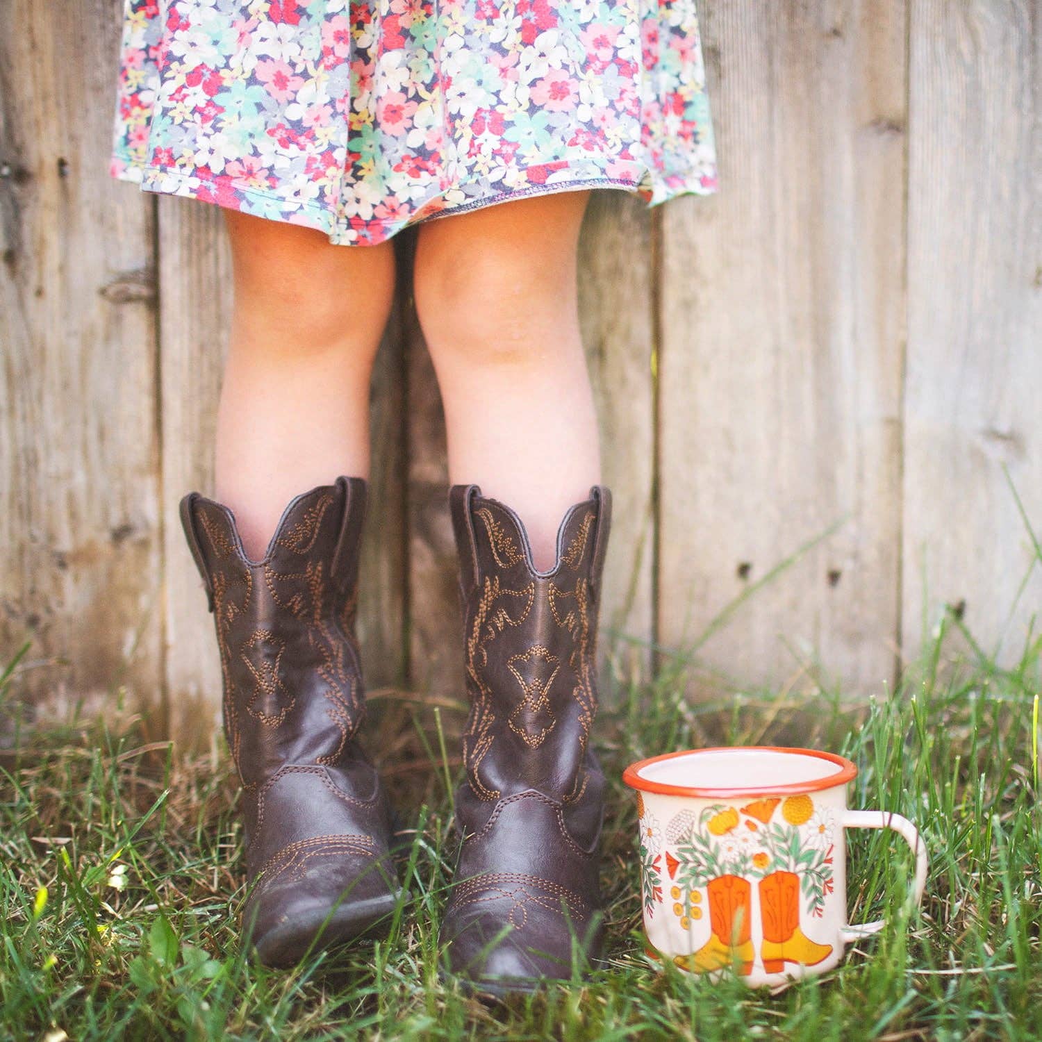 Person wearing cowboy boots and a floral dress standing in grass next to a mug with a wooden fence background.