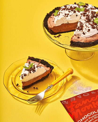 Chocolate pudding pie shown whole on a glass cake stand and a single slice on a glass plate with a fork, set on a bright yellow background with a red package corner visible.