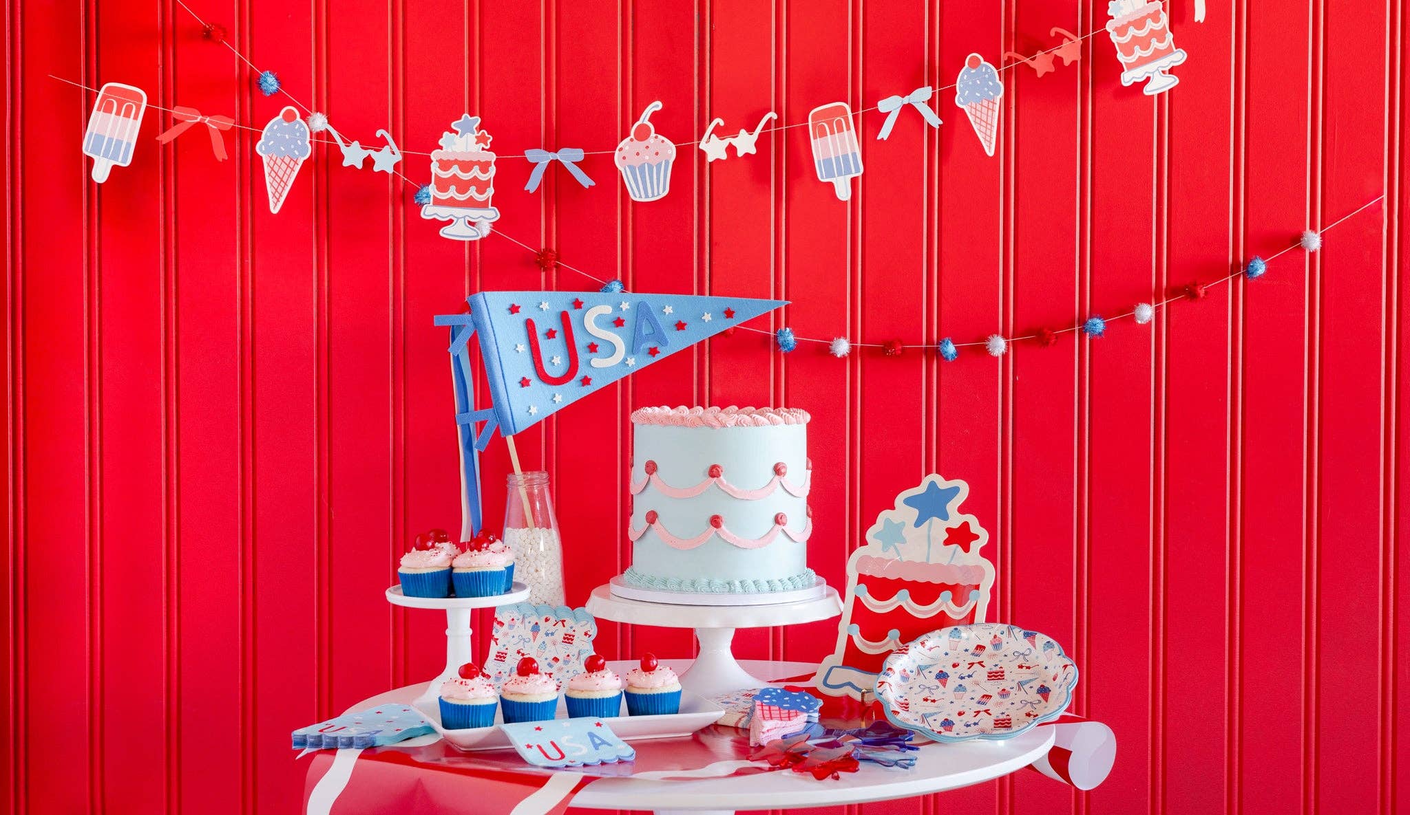 Decorative setup with a cake, cupcakes, and party decorations against a red background.