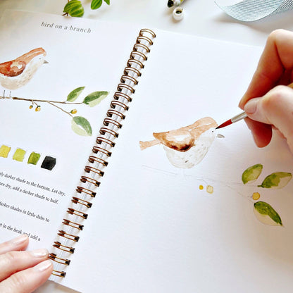 Person painting a bird illustration in a notebook with a paintbrush.