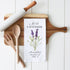 Wooden cutting board with rolling pin, wooden utensils, and a lavender-themed towel on a white surface.