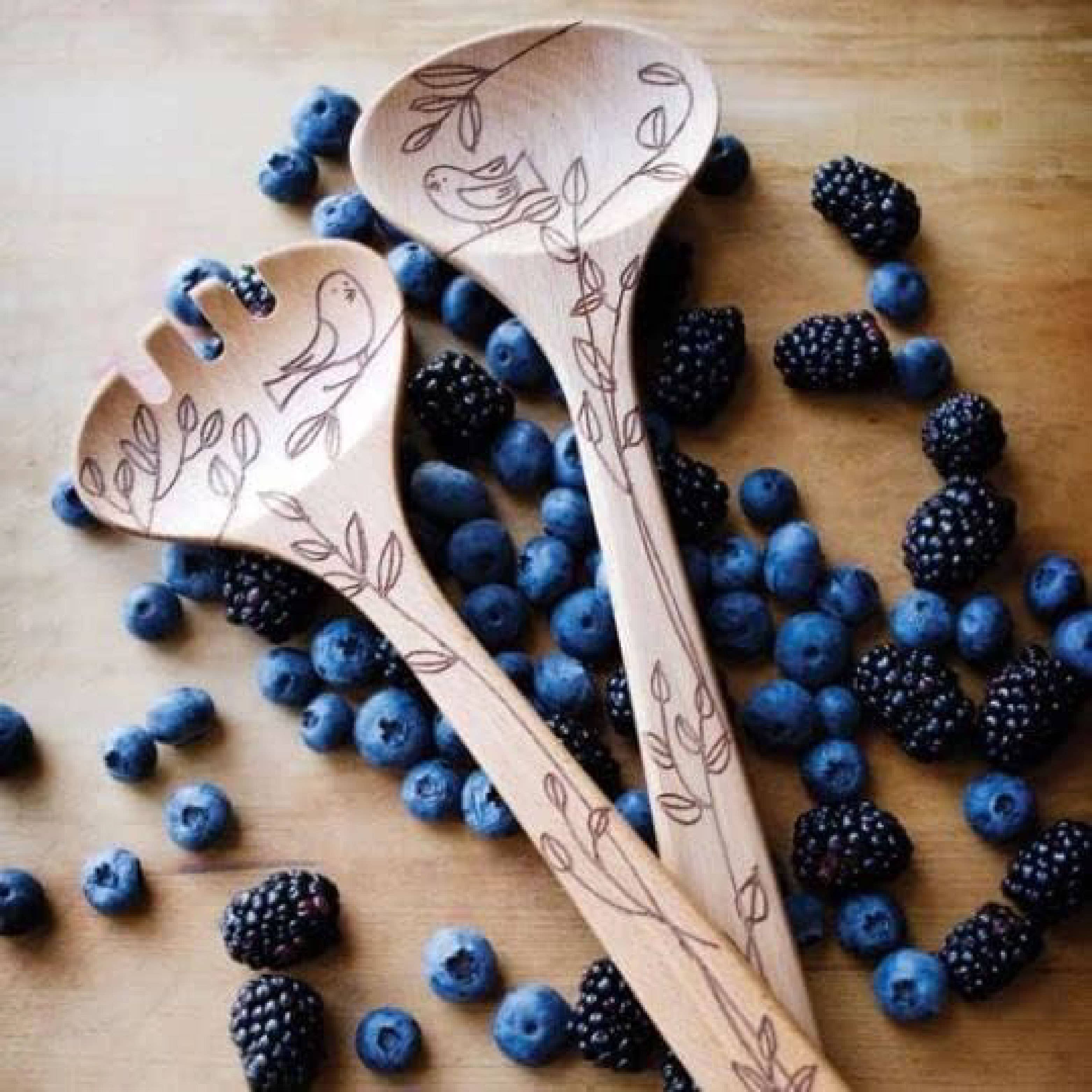 Wooden salad servers with engraved designs on a wooden surface with blueberries and blackberries.