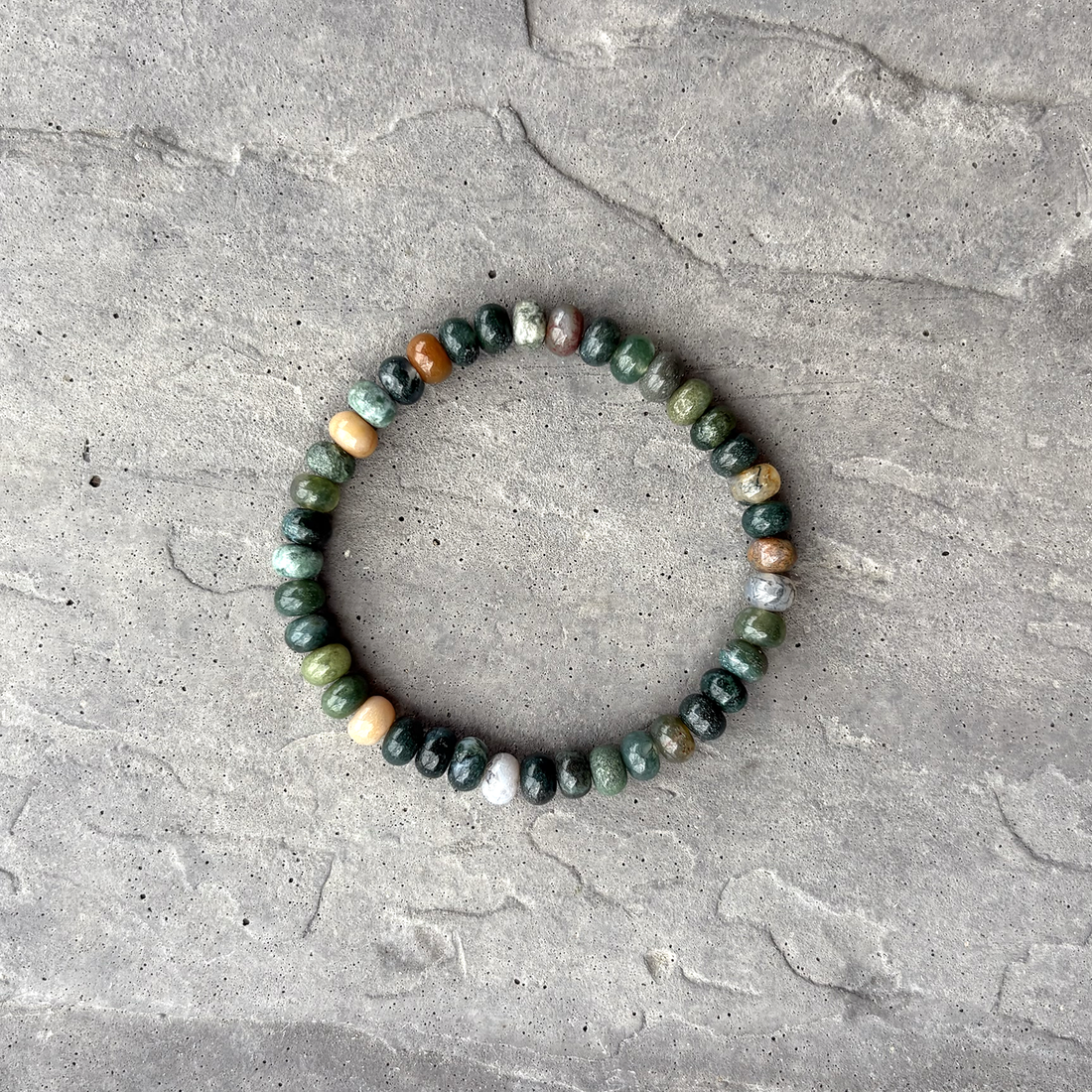 A beaded bracelet with round Indian Agate beads in shades of green, brown, and neutral colors, displayed on a textured grey background.