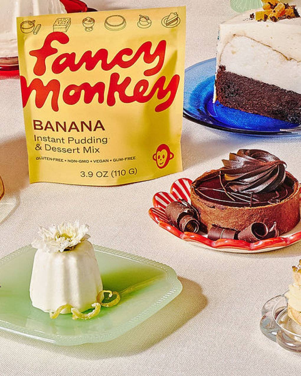 A bag of Fancy Monkey Banana Instant Pudding &amp; Dessert Mix stands on a table next to plated desserts, including a banana pudding, a chocolate cake, and a chocolate tart.