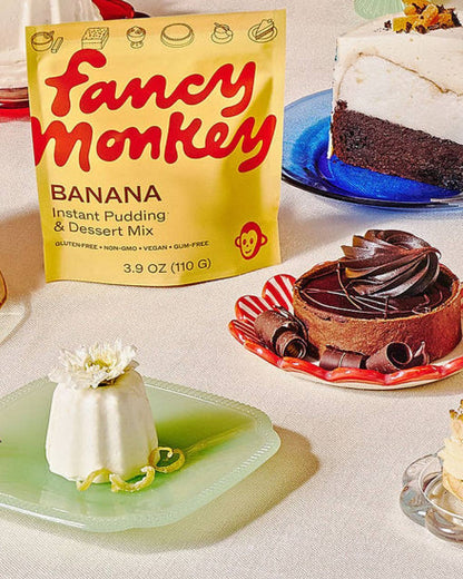 A bag of Fancy Monkey Banana Instant Pudding &amp; Dessert Mix stands on a table next to plated desserts, including a banana pudding, a chocolate cake, and a chocolate tart.
