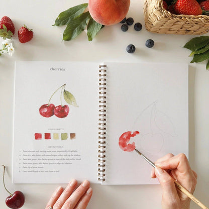 Person painting cherries in a sketchbook with fruits and flowers on a table