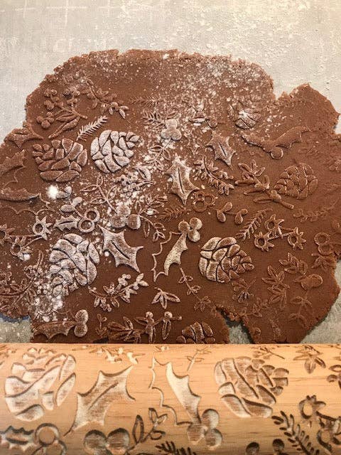 A decorative wooden rolling pin with carved botanical and holly leaf patterns rests on top of a rolled sheet of brown dough dusted with flour, set against a neutral gray background.