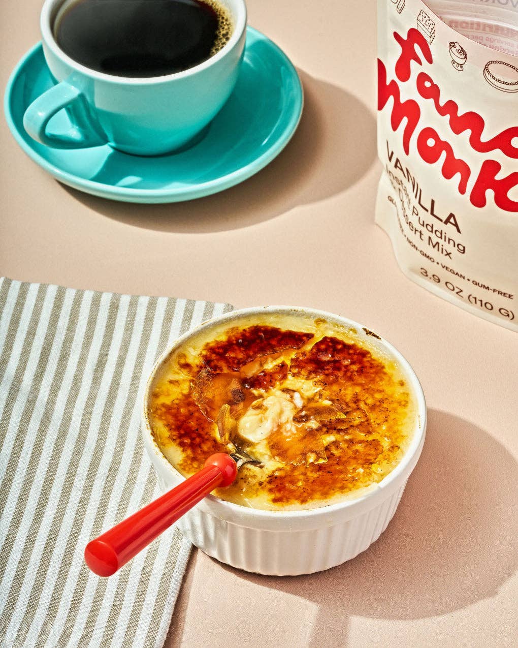 A bowl of crème brûlée with a caramelized crust sits on a striped napkin with a red spoon, next to a cup of black coffee and a bag of Fanu Monk Vanilla Pudding.