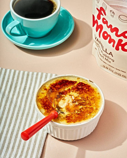 A bowl of crème brûlée with a caramelized crust sits on a striped napkin with a red spoon, next to a cup of black coffee and a bag of Fanu Monk Vanilla Pudding.