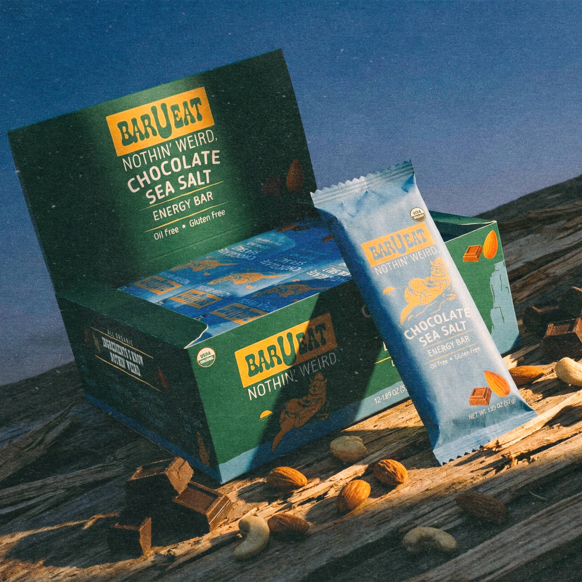 BAR U EAT Nothing Weird. Chocolate Sea Salt Energy Bar snack boxes and individual wrapped bars displayed on a rustic wooden surface with scattered chocolate pieces, nuts, and cashews against a blue sky background.