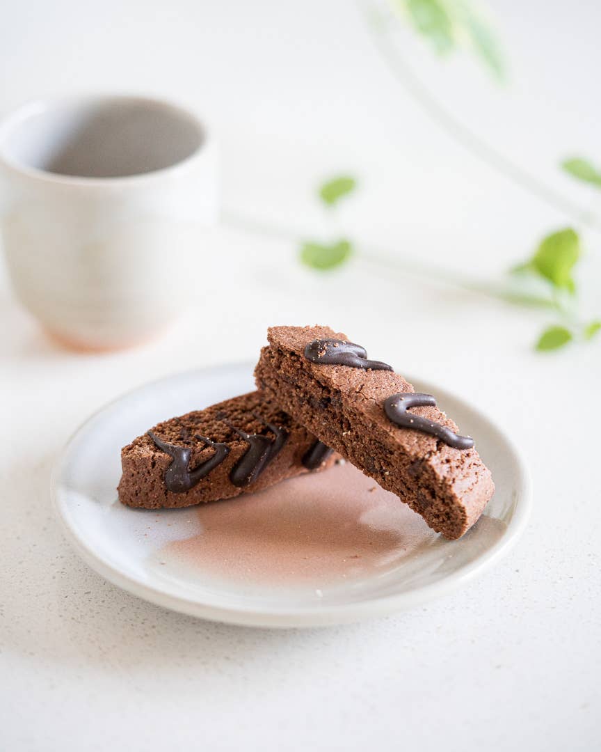 Two chocolate biscotti on a white plate with a blurred coffee cup in the background.
