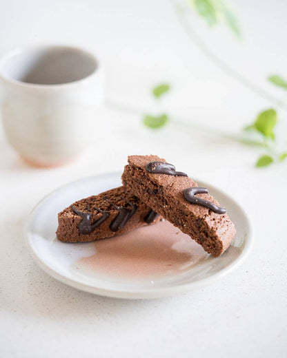 Two chocolate biscotti on a white plate with a blurred coffee cup in the background.
