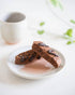 Two chocolate biscotti on a white plate with a blurred coffee cup in the background.
