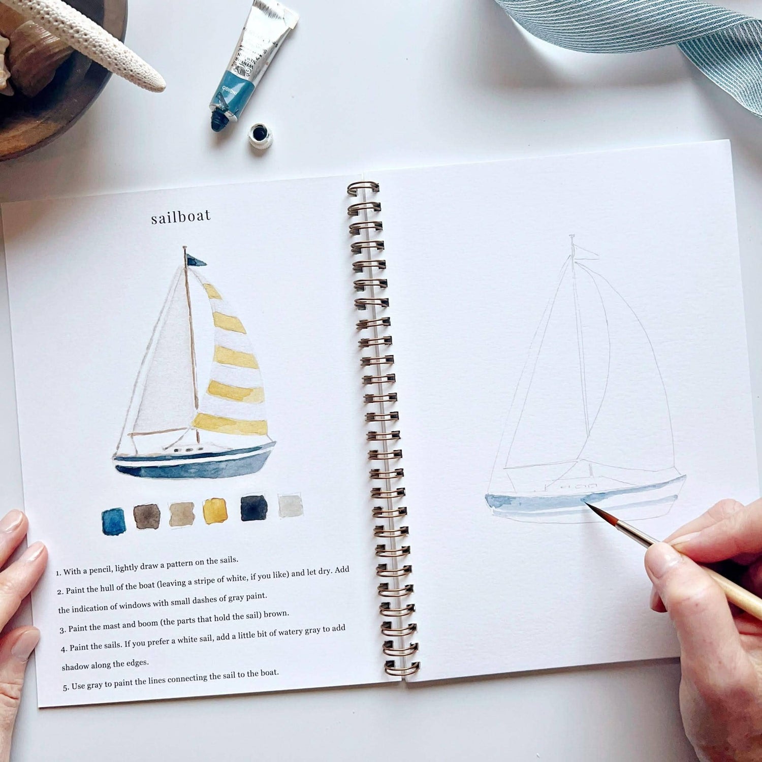 Open book with sailboat illustration and color chart, hands holding a paintbrush, on a white surface.