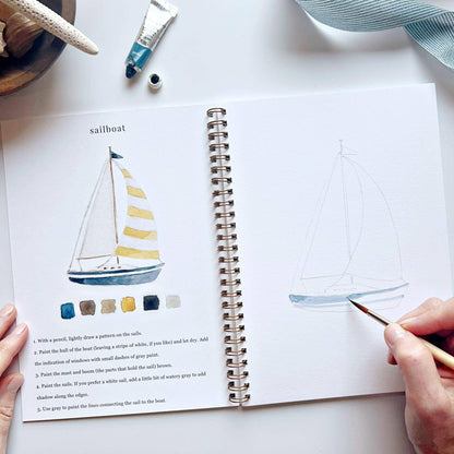 Open book with sailboat illustration and color chart, hands holding a paintbrush, on a white surface.