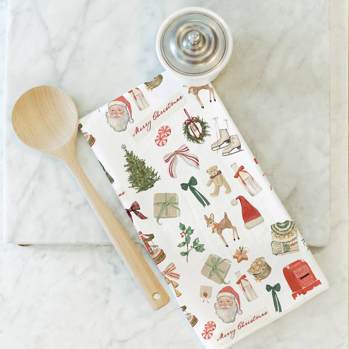 A flat-lay of a white kitchen tea towel with a colorful Christmas print, styled with a wooden spoon and a metal coffee grinder on a white marble surface.