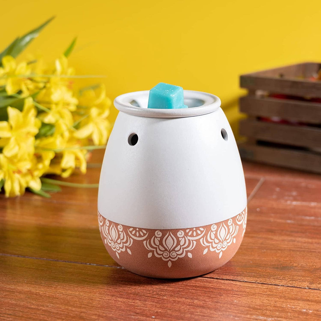 White and terracotta decorative wax warmer with a blue wax tart on a wooden surface with yellow flowers in the background.