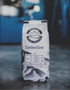 A bag of Silver Mountain Coffee Roasters Timberline light roast coffee stands on a dark surface with a blurred blue workshop background.